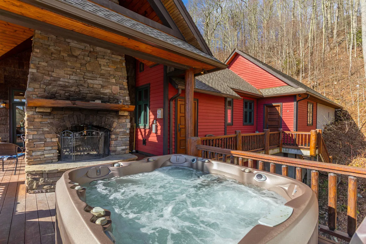 An inviting shot of a wooden deck featuring a bubbling hot tub, a stone fireplace, and cozy seating, all overlooking tranquil forest views. The setting is perfect for relaxing after a day of mountain adventures.