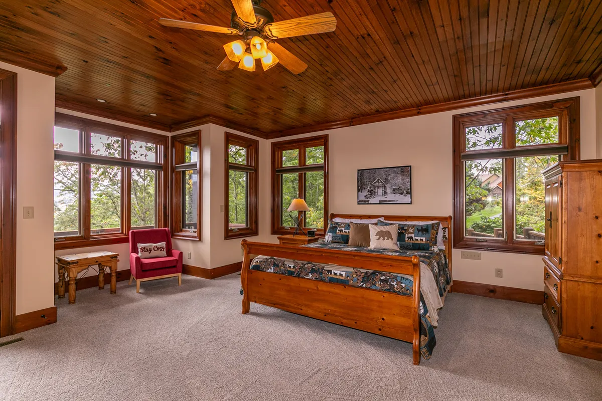 A spacious king bedroom featuring a large wooden bed, expansive windows that fill the room with natural light, and an inviting accent chair. The serene setting is ideal for relaxation, offering views of the surrounding trees and a comfortable atmosphere for reading or unwinding.