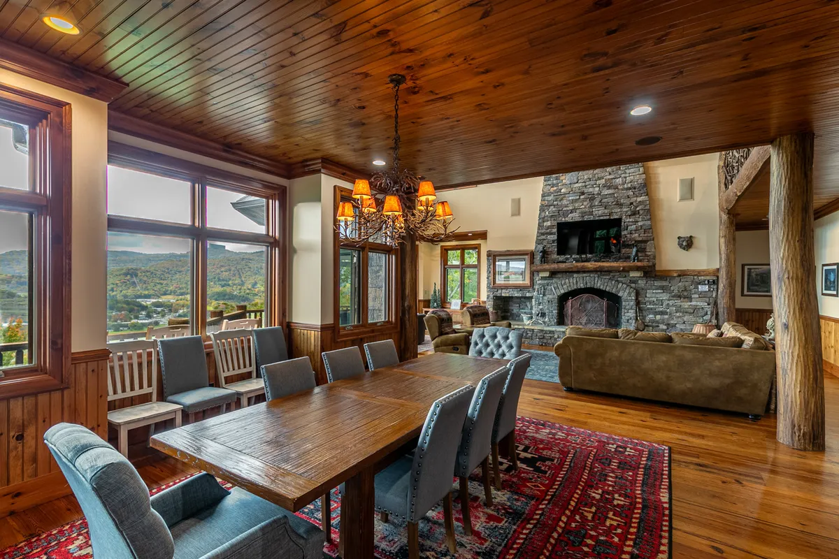 A dining area with a large wooden table set near a stone fireplace and a grand chandelier. Tall windows reveal serene views, and the ambiance suggests warm gatherings, game nights, and holiday celebrations.