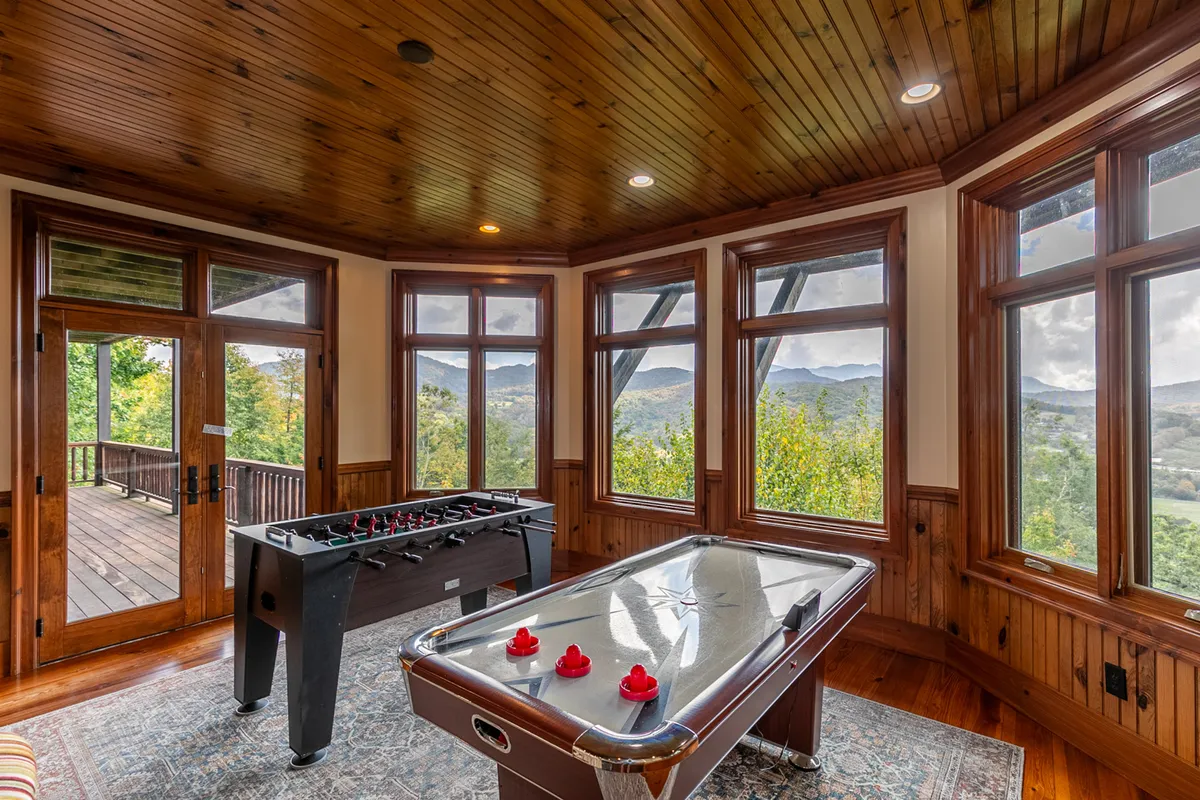 A lively secondary game room showcasing a foosball table and an air hockey table set in front of large windows, with wooden ceilings adding warmth and character. The room exudes a fun, laid-back vibe ideal for family competitions and group activities.