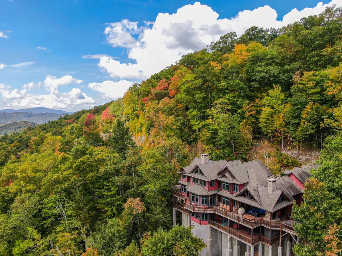 A wide exterior shot of the Overlook Estate, showcasing the large wooden multi-story house nestled among trees with a mountain backdrop. The house's spaciousness and connection to nature are evident, hinting at ample room for large families to gather and explore the area.