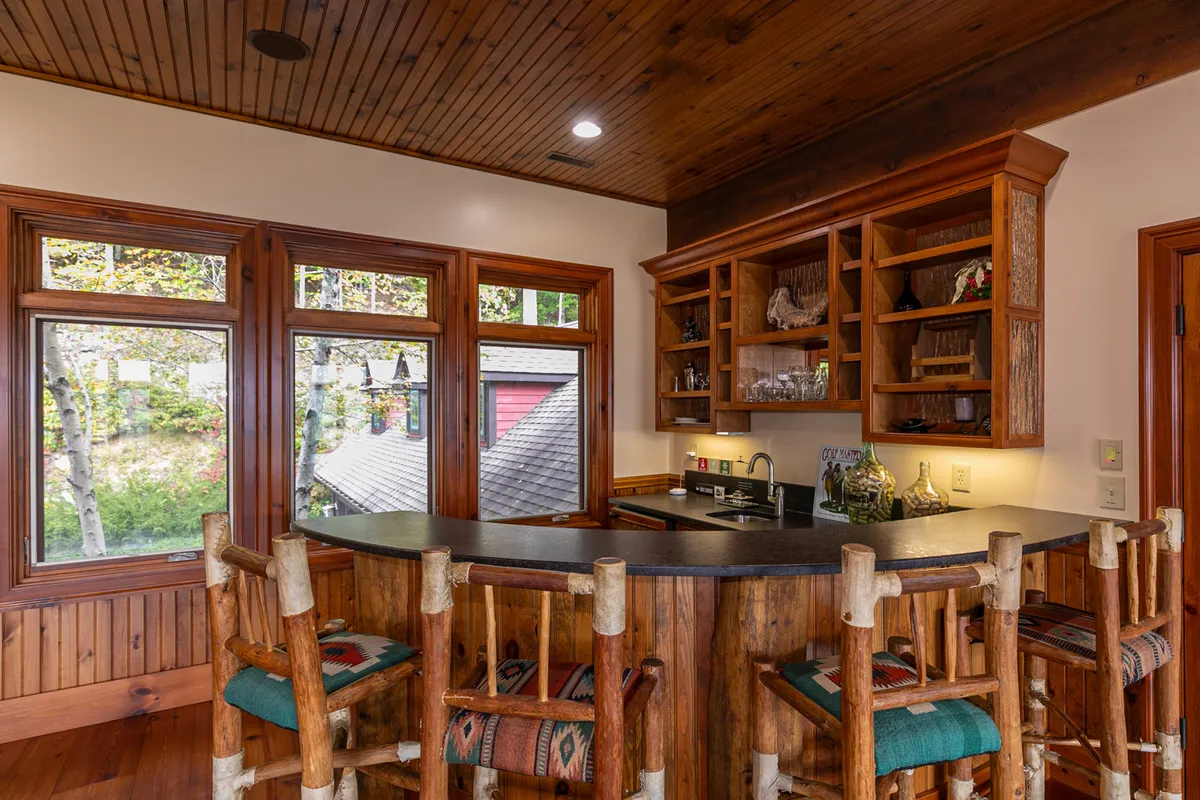 A well-appointed wet bar area with a wooden bar counter and upholstered stools, shelves stocked with glassware, and large windows providing a bright, inviting setting for mixing drinks and socializing.