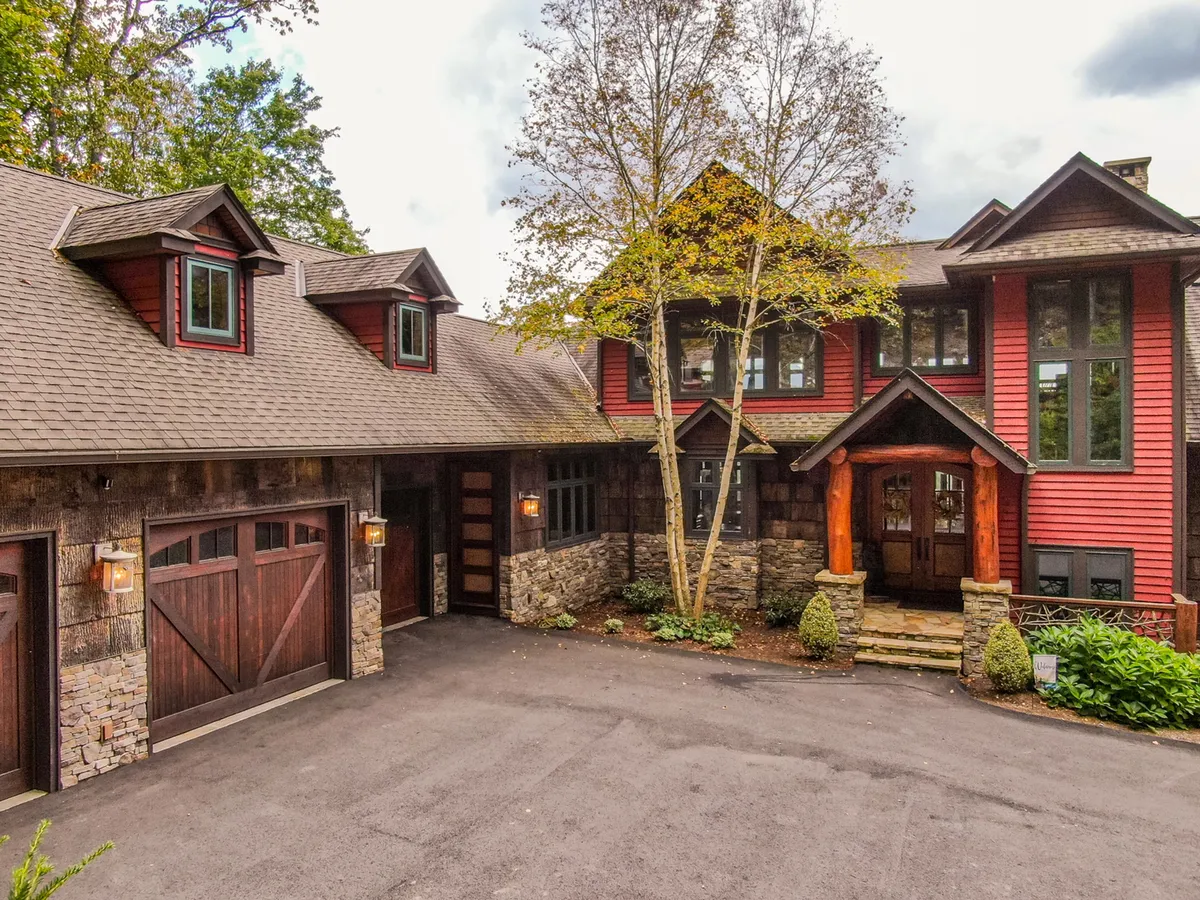 The main entrance of the estate, featuring warm wooden accents, a handsome stone facade, large windows, and mature trees that envelop the property, creating a sense of grandeur and welcome. The view hints at the tranquility and privacy of a luxury mountain retreat.