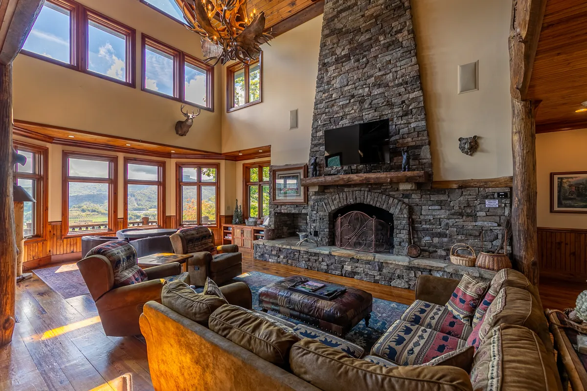 A great room featuring a dramatic stone fireplace, plush and comfortable seating, an antler chandelier overhead, and expansive windows revealing sweeping mountain views. The arrangement is perfect for gatherings, with ample space for relaxing by the fire or enjoying the scenery outside.