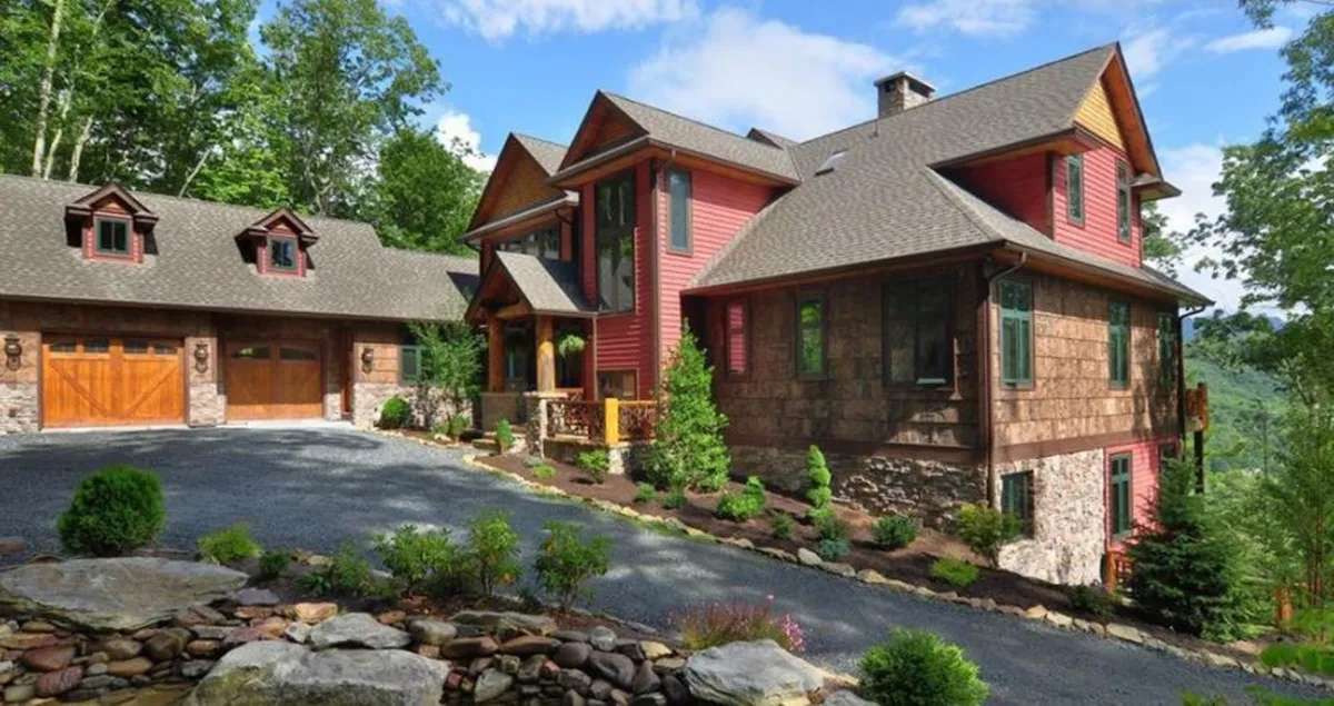 The image showcases the impressive main house and garage set amidst a beautifully landscaped garden, with a spacious porch perfect for gatherings. The serene mountain backdrop and lush surroundings create an inviting and peaceful atmosphere, ideal for family getaways and enjoying the outdoors.