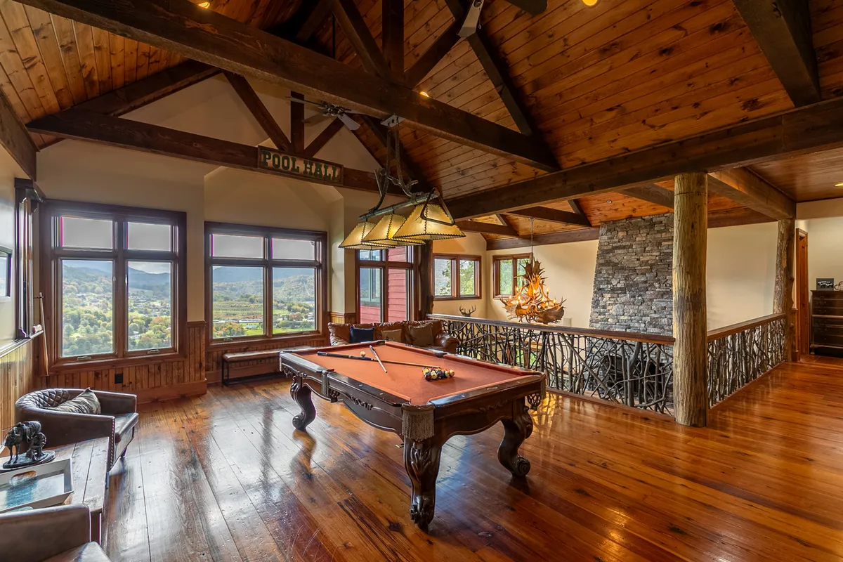 A spacious entertainment loft featuring a pool table as the centerpiece, surrounded by comfortable seating, rustic wooden beams overhead, and a stone fireplace. Large windows flood the space with natural light and offer scenic views, creating a welcoming area for games and social gatherings.